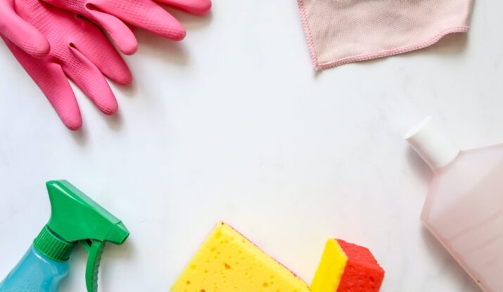 A flat lay of cleaning supplies on a white surface, including pink rubber gloves, a green spray bottle, a pink microfiber cloth, and two sponges, one yellow and one red.