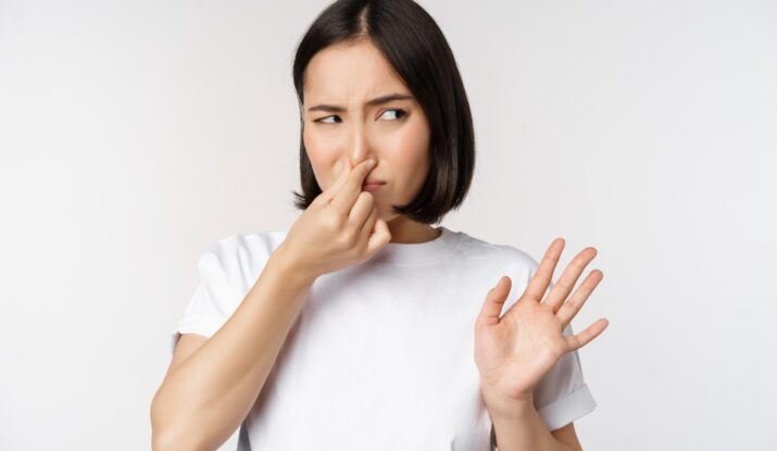 A woman in a white t-shirt is making a displeased face and holding her nose with one hand, indicating a bad smell, while gesturing stop with her other hand.