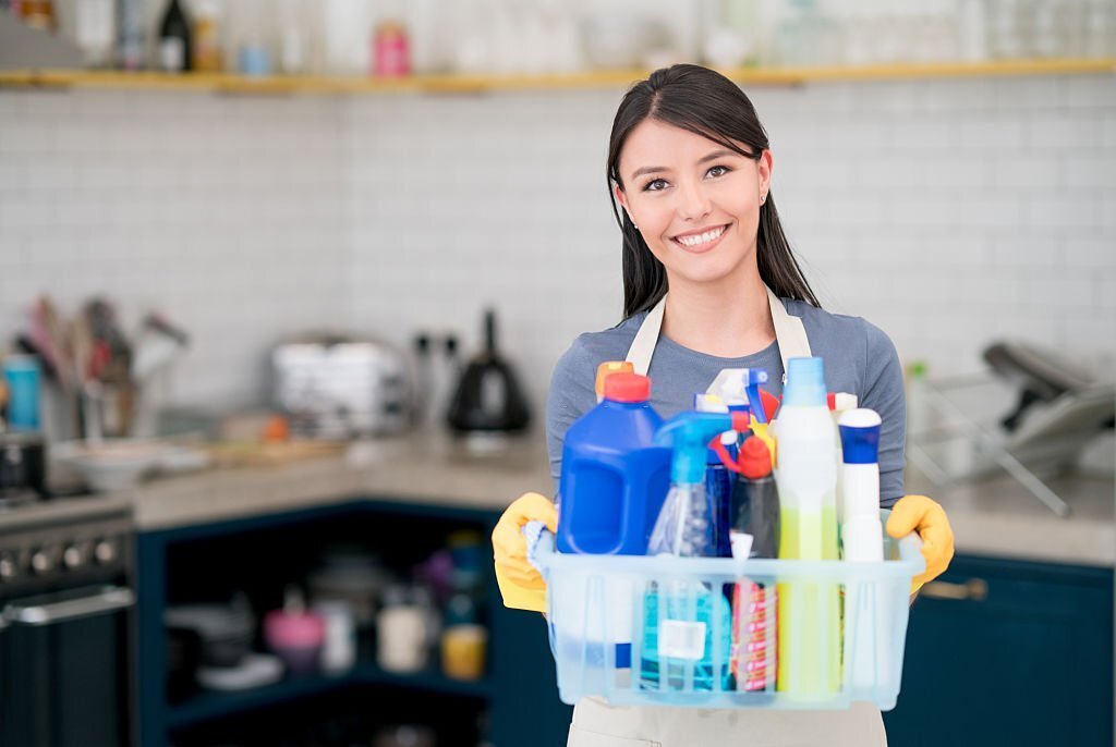 A woman in a grey t-shirt and white apron, wearing yellow gloves, holds a caddy of cleaning supplies with a kitchen in the background.