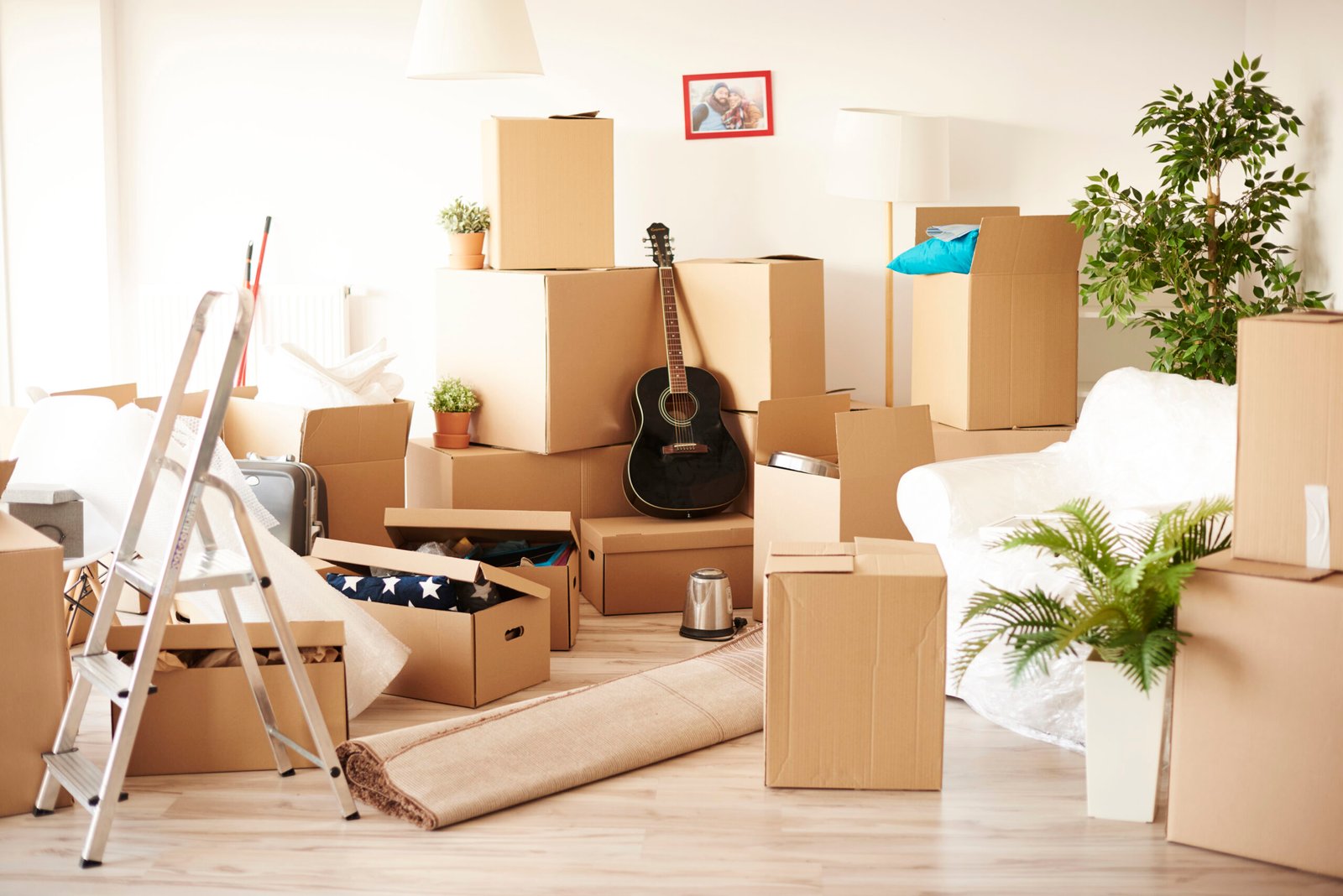 A room filled with unpacked moving boxes, various household items, and a rolled-up rug, indicating a moving day scene with belongings yet to be organized and set up in a new home